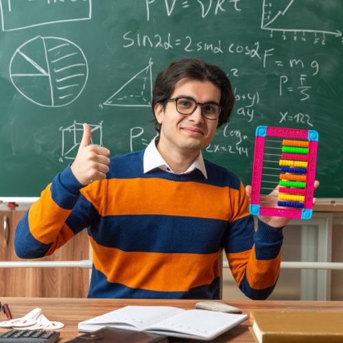 pleased-young-caucasian-geometry-teacher-wearing-glasses-sitting-desk-with-school-supplies-classroom-showing-abacus-thumb-up-looking-front Physics Tuition near me Class 11